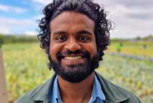 This image shows a dark brown man with curly hair and a beard, smiling broadly. He is wearing a blue shirt under a green jacket, and the background features a blurred outdoor setting with greenery and a partly cloudy sky.