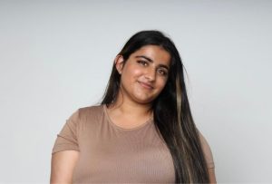 A picture of a Indian woman with long brown with blonde highlights hair with a white background with her head tilted to the right side and wearing a brown top and smiling.