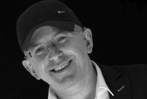 A man in a white shirt, dark jacket, cap with hearing aid smiling stood in front of a dark background.