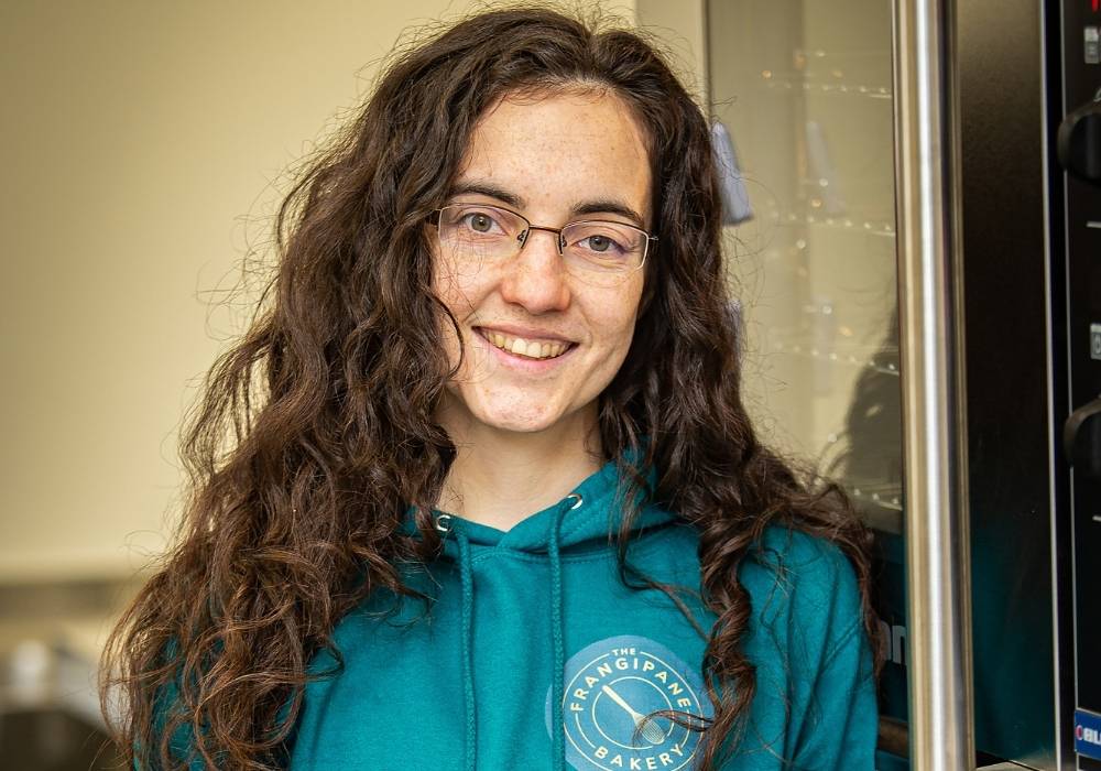 "Rachel, a white woman with long brown curly hair and glasses wears a teal hoodie with The Frangipane Bakery’s gold whisk logo on the left breast as she smiles in front of a commercial oven. Image credit: Jason Baxter, South of Scotland Enterprise"