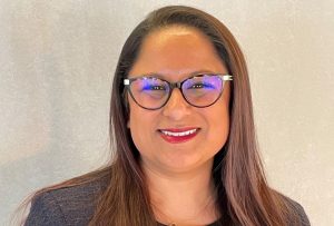 Reena is a woman of South Asian (Indian) heritage. She has long brown hair, brown eyes and brown skin. She is wearing tortoiseshell spectacles and a navy blue blazer with gold buttons. She is standing in front of a cream backdrop.