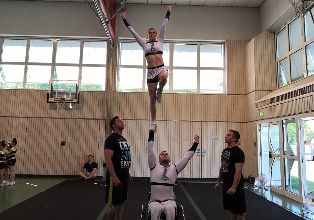 "A wheelchair user in a white and blue tight cheerleading uniform holds a woman in a matching uniform above his head in one hand, the woman is standing on one leg and has her arms stretched to the sky. Two men look on protectively one from each side ready to support if something goes wrong."