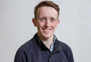 A brown haired white man in a navy blue jumper and light grey polo shirt stood in front of a grey background.