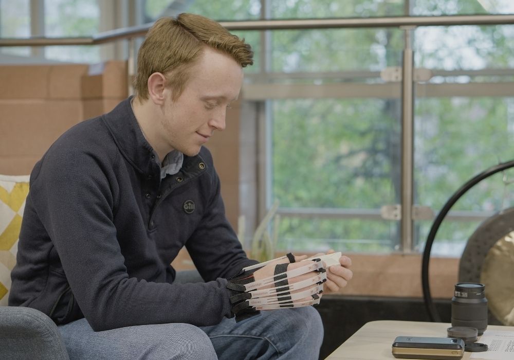 Robert a young white male adult with brown/ginger hair wearing a navy blue jumper with light blue jeans is sat down in a blue fabric chair whilst holding a white 3D print prototype with a dexterity simulation glove on his right hand. This is within an office space with railings and large windows behind Robert as well as a large metal gong and wooden table in front of him. - Credit - Ziggy Lyons