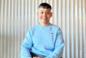 A wheelchair user with black hair, wearing a light blue jumper, and sat in front of a white wall.