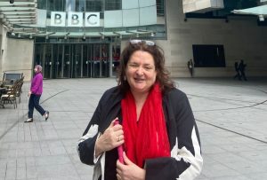 A dark haired woman who is smiling. She is dressed in a black jacket and vivid red scarf holding her white cane. She is standing in front of New Broadcasting House at the BBC.