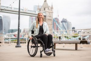 A blonde haired wheelchair user is wearing a black outfit with light blue denim jacket. She is sitting in her black manual wheelchair, smiling at the camera with one hand on the wheel and the other on her lap. The backdrop is Tower Bridge in London, and it is a light cloud day.