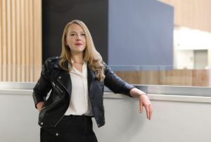 ginger woman with long hair, white shirt and black jacket leaning against a mid-level wall