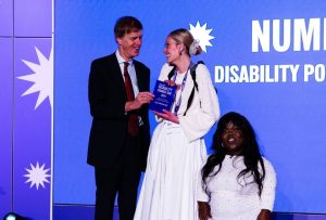 An image of Stephen Timms MP presenting Celia with her award. Celia is a white woman with dark blonde hair which is tied back into a bun. She is wearing a long white sleeved jumper and long white skirt. Next to her is Fats Timbo, a black woman of short stature wearing a white dress.