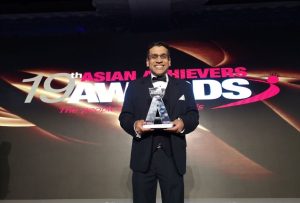 A dark haired Asian man, wearing a black tuxedo with a white shirt, holding the Asian Achievers Award for Entrepreneur of the Year in 2019.