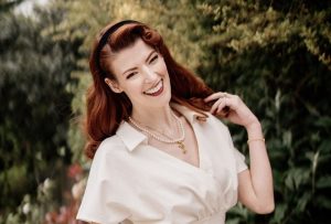 In her garden, Jessica, a white young woman, smiles broadly at the camera whilst one hand tosses her red hair over her shoulder. She is standing in front of a large, textured green bush that fills the background behind her. Her vibe is very vintage with pin-curled hair, a velvet headband and a 1950s cut silk blouse.