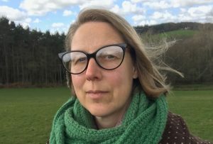 head and shoulders of a white woman with dark blond hair and glasses, wearing a wide green scarf and brown coat, standing outside in the countryside