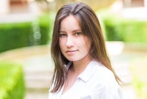 A young white person with long brown hair standing in front of a fountain. They have a loose white blouse, and dark trousers. They are looking directly at the camera with a half smile.