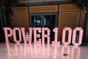 An image of large purple light up letters, which are standing from the floor. They spell Power 100.