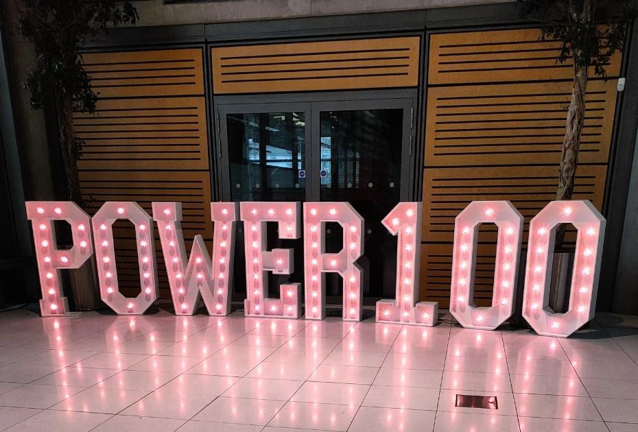 An image of large purple light up letters, which are standing from the floor. They spell Power 100.