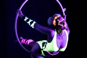 In a uv photoshoot, Rachael is lit by a violet purple light, she is posing in an aerial hoop. She is wearing a neon green costume with neon pink and green wraps around her forearm and calf. Her hair is in big space buns with neon orange highlights. Her make up is neon green. She is leaning through the front of the hoop, her hands grip the hoop behind her. He back leg is horizontal to the ground, her front leg is bent; toes pointed.